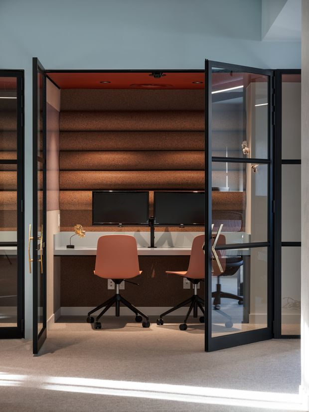 Compact enclosed home office or study nook with two workstations, dual monitors, terracotta upholstered task chairs, and a white desk. The space is framed by black metal-and-glass doors and features warm brown acoustic wall panelling, a matching textured ceiling, integrated lighting, and soft carpet flooring. A glass door reflection reveals additional office seating and reinforces the workspace setting.