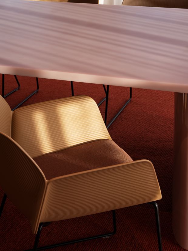 Close-up interior detail of a sculptural ribbed brown lounge chair with black metal legs, positioned on deep red carpet beneath a pale pink veined stone or solid-surface table. The image emphasizes tactile materials, warm tones, and contemporary furniture design.