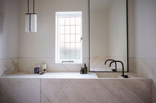 A refined bathroom vanity with a long pale stone countertop and matching full-height cabinetry or panelled fronts. The space features a black dual-basin faucet, large black-framed mirror, marble-look wall splashback, a tall frosted-glass window, and minimalist pendant lighting. Neutral warm tones and clean lines create a calm, understated contemporary setting.