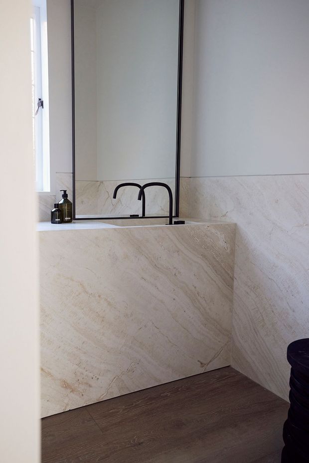 A refined bathroom vanity area featuring a large frameless mirror, double black wall-mounted faucets, and a floating vanity clad in heavily veined pale natural stone. Matching stone wall wainscoting continues around the room, contrasting with dark timber-look flooring. A narrow window and soap dispensers are visible beside the vanity.