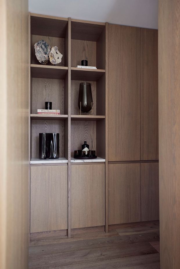 A close-up interior view of a custom built-in timber display and storage wall. Open shelving contains decorative objects, books and glassware, while closed lower and side cupboards provide concealed storage. The joinery and surrounding walls are finished in warm medium-to-dark wood tones, with a matching timber floor and a restrained, refined styling.