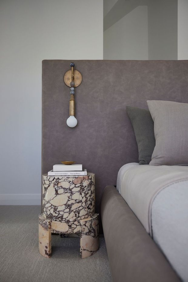 Close-up of a refined bedroom corner featuring a bed with a taupe upholstered headboard and matching upholstered bed base, layered neutral pillows and bedding, a sculptural brass-and-globe wall light, and a distinctive cylindrical stone side table with dramatic veining. The room has pale grey walls, a textured grey carpet, and a muted, luxurious neutral palette.