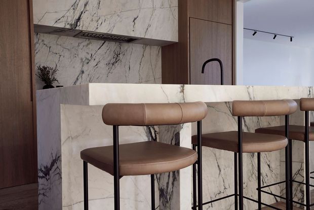 A refined contemporary kitchen with a marble-clad island and matching marble splashback, integrated dark timber cabinetry, and a curved black tap. Several tan leather bar stools with slim black metal frames line the island. The space has a warm, luxurious palette and clean linear detailing.