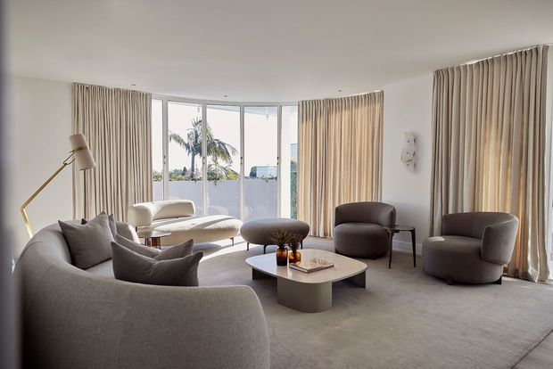 A bright, contemporary living room with a large curved sectional sofa, rounded lounge chairs, a low sculptural coffee table, and a slim floor lamp. Full-height curved glazing and layered beige curtains open to a tropical exterior view, while soft neutral upholstery and carpet create a calm, refined atmosphere.