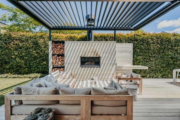 An outdoor entertaining area with a large modular lounge arranged around a built-in fireplace, sheltered by a slatted pergola. The space features timber decking, a concrete or rendered fireplace wall with integrated log storage, a hedge backdrop, and abundant natural light with strong linear shadows.