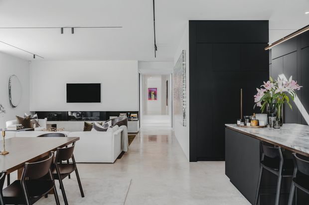 Bright open-plan contemporary interior showing a kitchen, dining area and living room. The kitchen features matte black full-height cabinetry, a marble-look benchtop and integrated island seating. A white sectional sofa faces a wall-mounted television, while a dining table with dark chairs sits in the foreground. Pale polished flooring, white walls and ceiling, minimalist linear lighting, and black accents create a clean, high-contrast scheme.