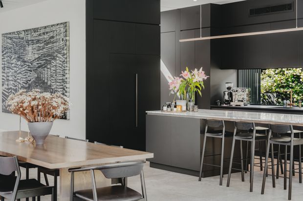 A sophisticated open-plan kitchen and dining area with a dark, minimalist kitchen featuring full-height black cabinetry, a long stone island with bar seating, integrated appliances, and a contemporary linear pendant light. A timber dining table with modern upholstered chairs occupies the foreground, complemented by monochrome wall art and floral arrangements. The space has a restrained black, grey, white, and timber palette with abundant natural light.