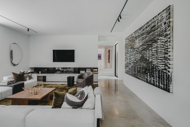 A spacious, contemporary living room with a large white sectional sofa, dark leather armchair, timber coffee table, and low black media cabinetry beneath a wall-mounted television. White walls and ceiling are accented by black track lighting, a circular wall mirror, and a large monochrome abstract artwork. The polished concrete floor and open hallway contribute to a clean, refined minimalist aesthetic.