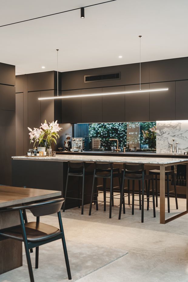 A sophisticated dark-toned kitchen and dining space with full-height charcoal cabinetry, a long marble-look island with integrated seating, black bar stools, and a linear pendant light. A mirrored or glazed backsplash reflects greenery and the kitchen work area, while a coffee station, sink, tapware, and floral arrangement add detail. The floor is a smooth light concrete finish, and a dining table with chairs occupies the foreground.