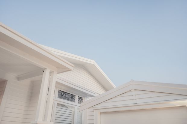 Close-up exterior architectural view of a predominantly white weatherboard home, showing overlapping pitched gable roofs, broad eaves, white timber posts, horizontal siding, and a detached or projecting garage-like gabled structure. The image is minimally styled and set against a clear pale blue sky.