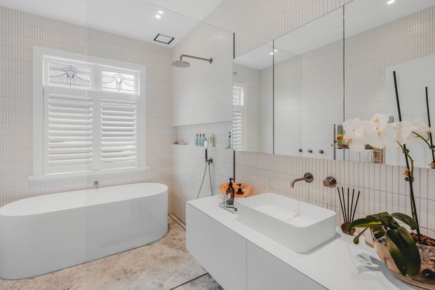 Bright, refined contemporary bathroom with a freestanding white bathtub beside a shuttered window, a wall-mounted vanity with white vessel basin, large mirrored wall, wall-mounted brushed metal tapware, and full-height narrow white tiled walls. Orchids and toiletries add small decorative accents. The room has a clean, airy, neutral palette and a standard-height ceiling.