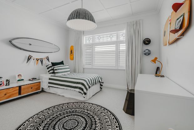 Bright, neatly styled bedroom with white walls, plantation shutters, full-height curtains, and pale carpet. A bed with layered green-and-white striped bedding sits beside a low timber-fronted cabinet, while a large black-and-white patterned round rug, surfboard wall decor, bunting, pendant light, and compact desk create a playful coastal feel.