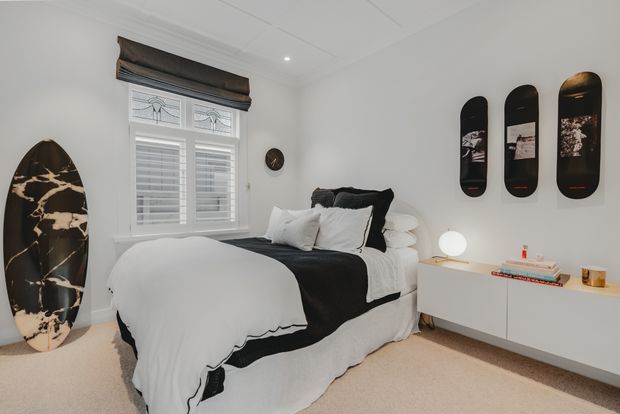 A bright, compact bedroom with white walls, white plantation shutters, and a neatly made bed dressed in white bedding with black accents. A black-and-white surfboard leans against the left wall, while three vertically mounted black boards provide graphic wall decor above a minimalist white floating cabinet. The room has carpet flooring, a small round lamp, and understated contemporary styling.
