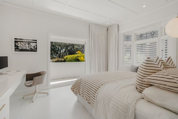 Bright, airy bedroom with a large upholstered bed dressed in layered beige and white striped bedding, white flooring and walls, full-height white curtains, plantation shutters, and decorative leadlight-style window panels. A compact white desk and modern upholstered chair sit beside the bed, with greenery visible outside the windows.
