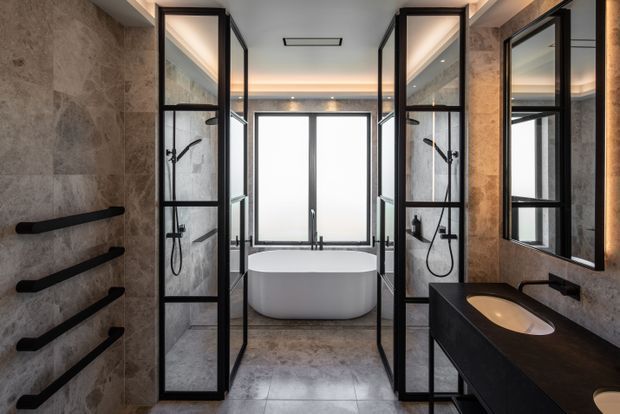 Luxurious contemporary bathroom with a freestanding white bathtub positioned in front of a large frosted window, flanked by two black-framed glass shower enclosures. The room features dark stone-look wall and floor finishes, black fixtures and towel rails, a dark floating vanity with an integrated basin, large black-framed mirror, and warm concealed lighting.