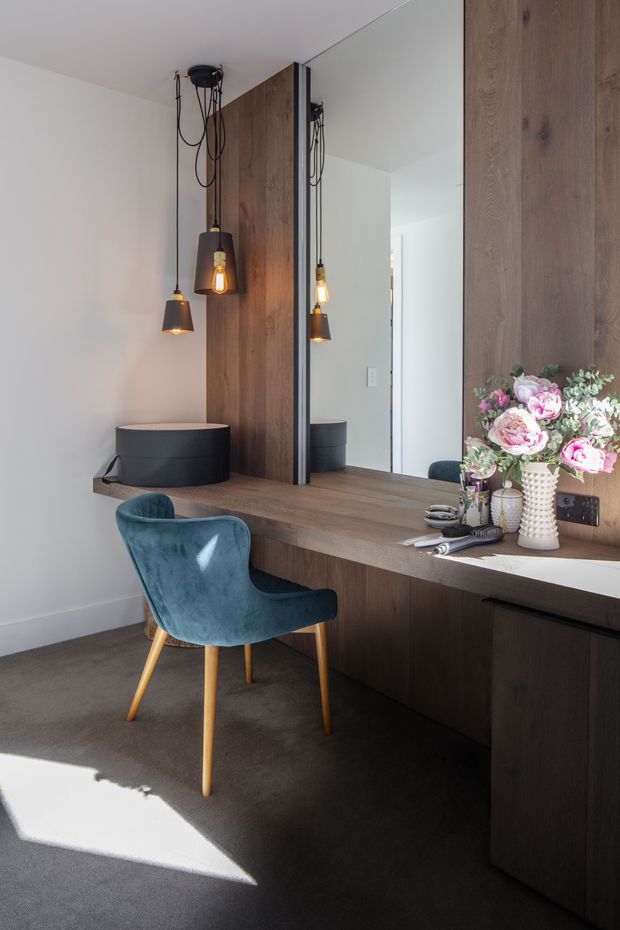 A compact, refined dressing or vanity area with dark timber built-in cabinetry and a long timber counter beneath a large mirror. Three black pendant lights hang over the counter, accompanied by a deep blue upholstered chair, ceramic vessels, and a floral arrangement. The room has white walls, dark carpet, and strong natural sunlight.