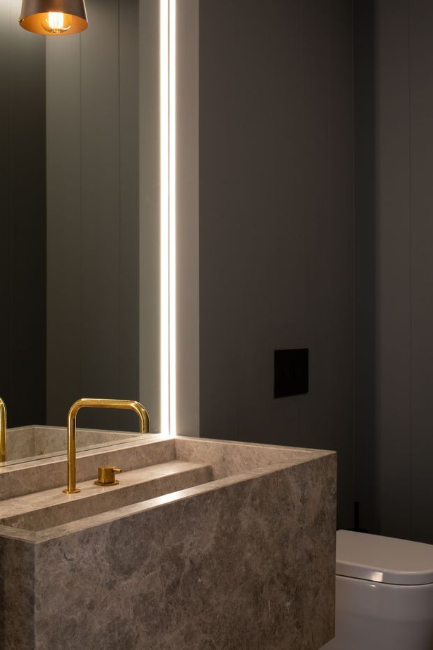 A dark, sophisticated bathroom featuring a monolithic stone vanity with integrated basin, a polished brass gooseneck faucet, large mirror with vertical integrated lighting, charcoal vertically panelled walls, a black wall switch, and a partially visible white toilet. A warm metallic pendant light adds contrast.