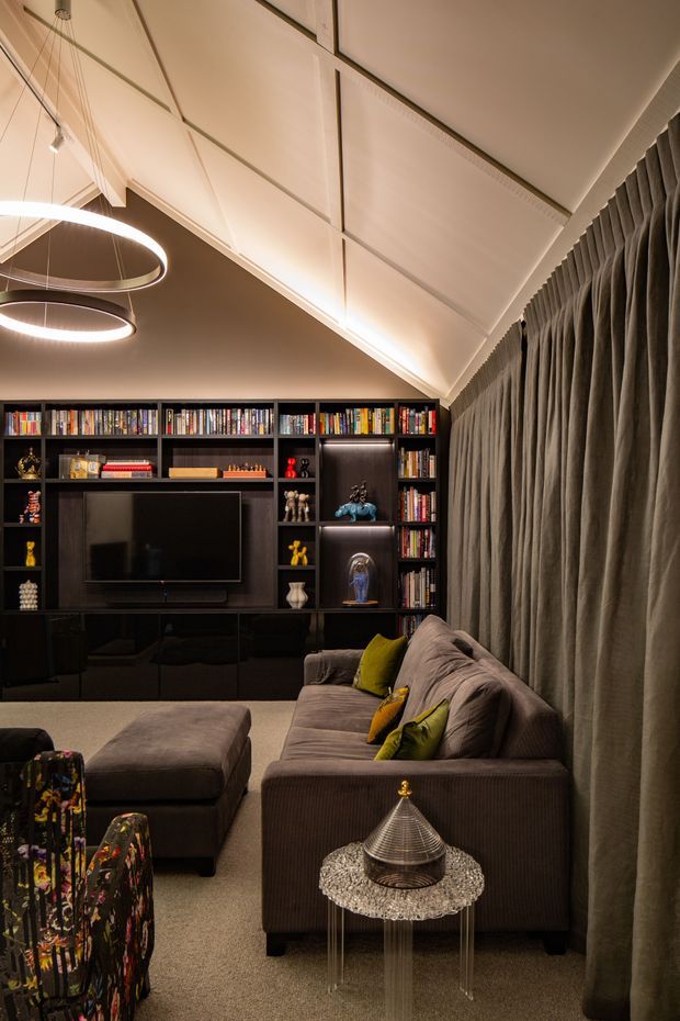 A sophisticated, moody media room with a large wall-mounted television integrated into dark built-in shelving filled with books and display objects. A substantial taupe sectional sofa, ottoman, patterned accent chair, green cushions, and a small decorative side table occupy the foreground. Floor-to-ceiling pleated curtains cover the right wall, while a dramatic pitched, paneled ceiling and suspended circular pendant lights add architectural interest.