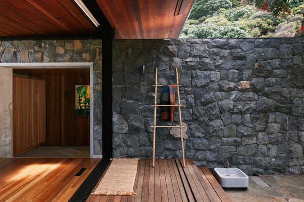 A sheltered outdoor courtyard or terrace adjoining a timber-lined interior, featuring a dark rough-stone masonry wall, timber ceiling and decking, a simple wooden ladder-style rack, woven mat, and a small white basin. Dense vegetation is visible above the wall, creating a natural, secluded setting.