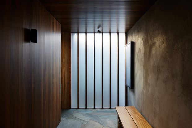 A dark, refined interior passage or entry area with vertical dark timber wall panelling on the left, a textured plaster or concrete-like wall on the right, and a matching timber ceiling. Tall translucent glazing with slim vertical timber mullions forms the end wall, bringing in diffused daylight. A narrow built-in timber bench runs along the right wall, with wall-mounted black fixtures or screens. The space has a minimalist, moody architectural character.
