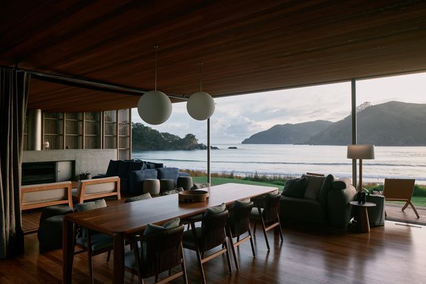 An open-plan coastal living and dining space with floor-to-ceiling glazing overlooking a sheltered bay and mountainous shoreline. The room features dark timber ceilings and flooring, a large timber dining table with upholstered chairs, a dark sectional sofa, lounge chairs, a built-in media or fireplace wall, and oversized spherical pendant lights. The interior has a warm, refined contemporary feel and strong indoor-outdoor connection.