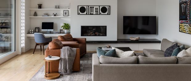 A contemporary living room with a large neutral sectional sofa, leather lounge chairs, a timber floor, built-in dark media cabinetry, wall-mounted television, fireplace, open shelving, and generous glazing with vertical blinds. The room includes a compact study nook and restrained modern artwork.