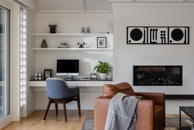 Refined contemporary living room and compact home office nook with built-in white shelving and desk, a computer monitor, sculptural decor, and a blue upholstered chair. A tan leather lounge chair with a grey throw occupies the foreground, while a linear electric fireplace and monochrome geometric artwork create a strong feature wall. The room has pale timber flooring, abundant daylight, and crisp white architectural detailing including a coffered ceiling and glazed doors.