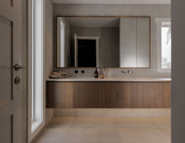 A refined contemporary bathroom vanity with a wall-mounted timber cabinet, pale stone or tile benchtop, large framed mirrored cabinet, dual wall-mounted basins and minimalist fittings. Large-format beige-grey tiles cover the walls and floor, with a window providing natural light.
