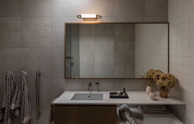 A refined contemporary bathroom vanity area with a large rectangular mirror, wall-mounted illuminated fixture, double-basin-style vanity counter, dark timber cabinetry, and grey tiled walls. Striped towels hang beside the vanity, while ceramic vessels and dried floral arrangements add warmth to the otherwise subdued, earthy scheme.
