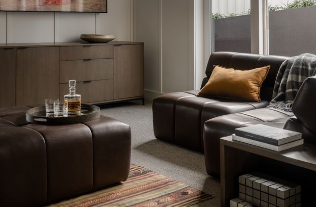 A refined living room with deep brown leather seating, a matching upholstered ottoman, a dark timber sideboard, and a black side table. A warm patterned rug, ochre cushion, monochrome throw, ceramic bowl, and books add texture and understated character. Large windows bring in natural light, with planter boxes visible outside.