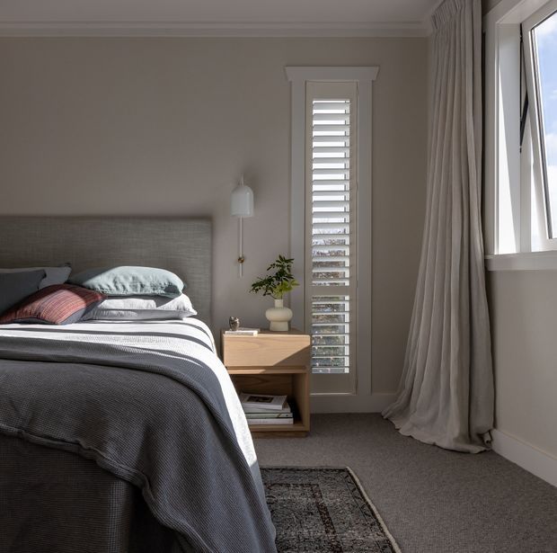 A softly lit, refined bedroom with a textured upholstered bedhead, layered grey bedding, a timber bedside table, pendant light, small plant, patterned rug, full-height curtains and white plantation shutters. The room has a calm neutral palette and traditional architectural detailing such as decorative cornices and framed window trim.