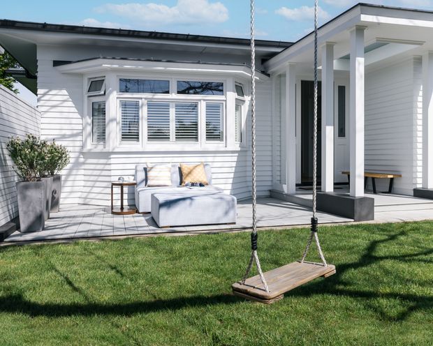Bright exterior view of a white weatherboard house with a projecting bay window, covered entry porch supported by square columns, and a small outdoor seating area on a paved terrace. A wooden swing hangs in the foreground over a manicured lawn.