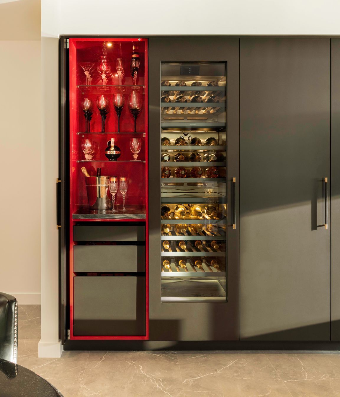 Beyond the integrated fridge, continues a freezer, wine chiller and hidden wine bar each embellished with black snake-skinned handles with polished brass-tip detailing; a nod to gold-leafed furniture collections.  Amongst this tall charcoal block, the displayed backlit wine collection is a welcome golden glowing interruption.