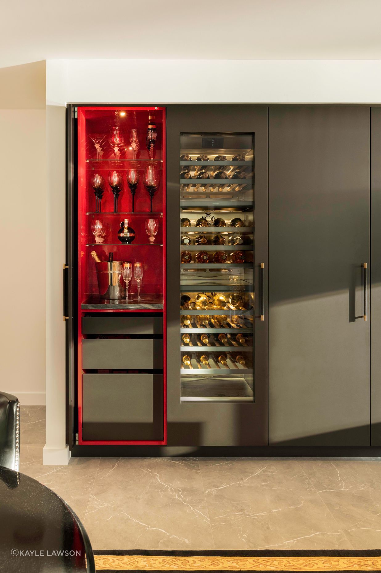 Beyond the integrated fridge, continues a freezer, wine chiller and hidden wine bar each embellished with black snake-skinned handles with polished brass-tip detailing; a nod to gold-leafed furniture collections. Amongst this tall charcoal block, the displayed backlit wine collection is a welcome golden glowing interruption.