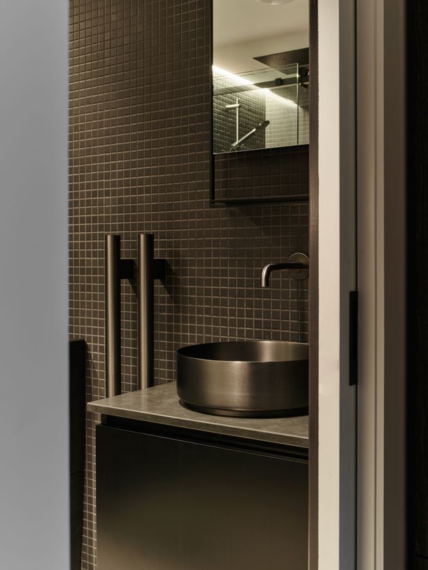 A compact, moody bathroom viewed through an open doorway, featuring dark small-format tiled walls, a floating dark vanity with a grey stone benchtop, a round brushed-metal vessel basin, dark wall-mounted tapware, and a large mirrored cabinet reflecting the shower area. Recessed and concealed lighting creates a refined, low-light atmosphere.