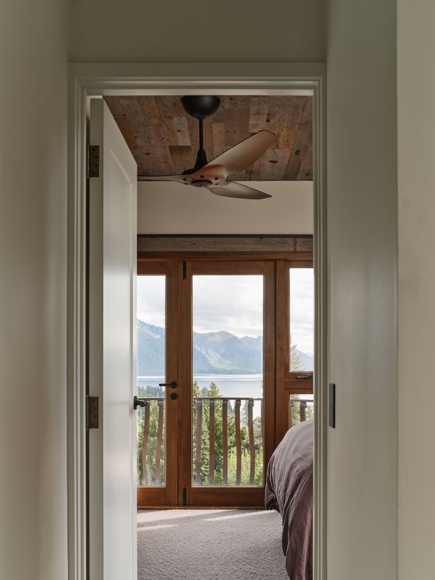 View through a doorway into a bedroom with a partially visible bed, carpeted floor, timber-framed glazed balcony doors, and a mountain-and-lake landscape beyond. The room features a reclaimed timber-plank ceiling, exposed dark timber trim, and a central ceiling fan, creating a warm natural alpine character.