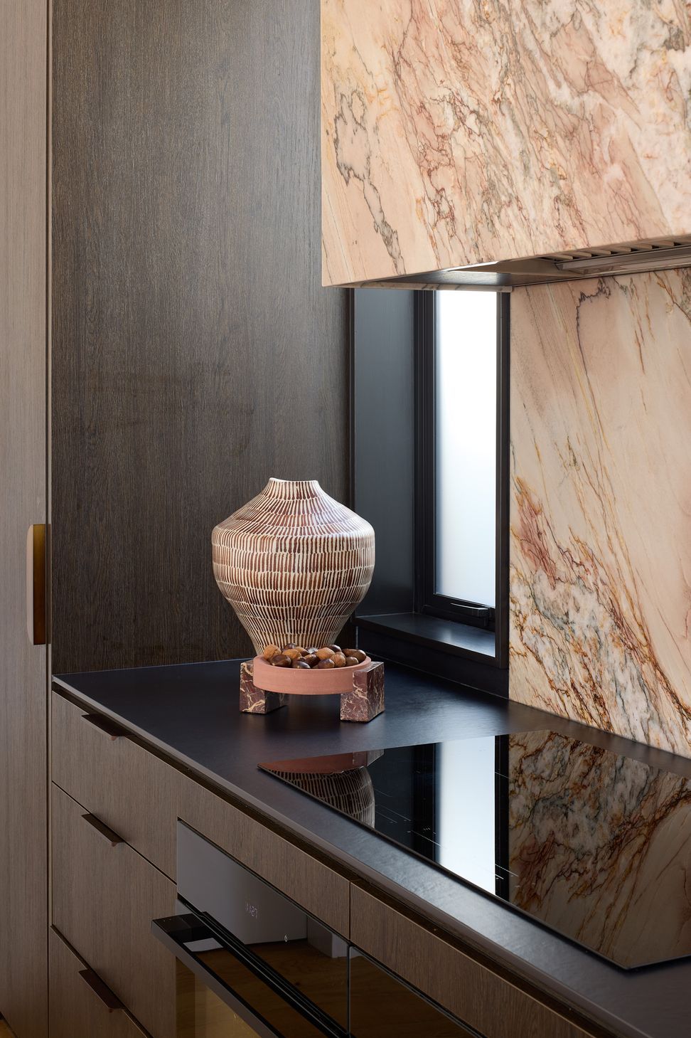 The rear kitchen bench pairs a dramatic granite splashback with dark oak cabinetry and a black Dekton worktop. Integrated appliances and refined detailing create a clean, contemporary workspace.