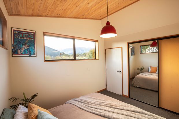 Warm, compact bedroom with a timber-lined ceiling, cream walls, dark carpet, and a low-profile bed dressed in neutral bedding. A red pendant light, framed artwork, indoor plant, and timber-framed horizontal window add character. A mirrored sliding wardrobe reflects an adjacent sleeping area or part of the room, while a white interior door opens beside it.