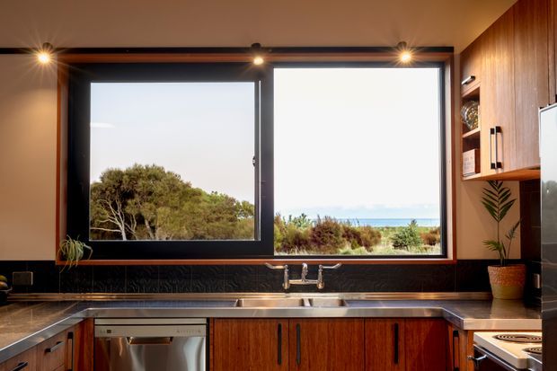 A warm, mid-century-inspired kitchen with dark-framed picture windows overlooking coastal vegetation and the ocean. The room features rich timber cabinetry, a dark textured backsplash, stainless-steel appliances and sink, stone-look benchtops, integrated display shelving, and warm spot lighting.
