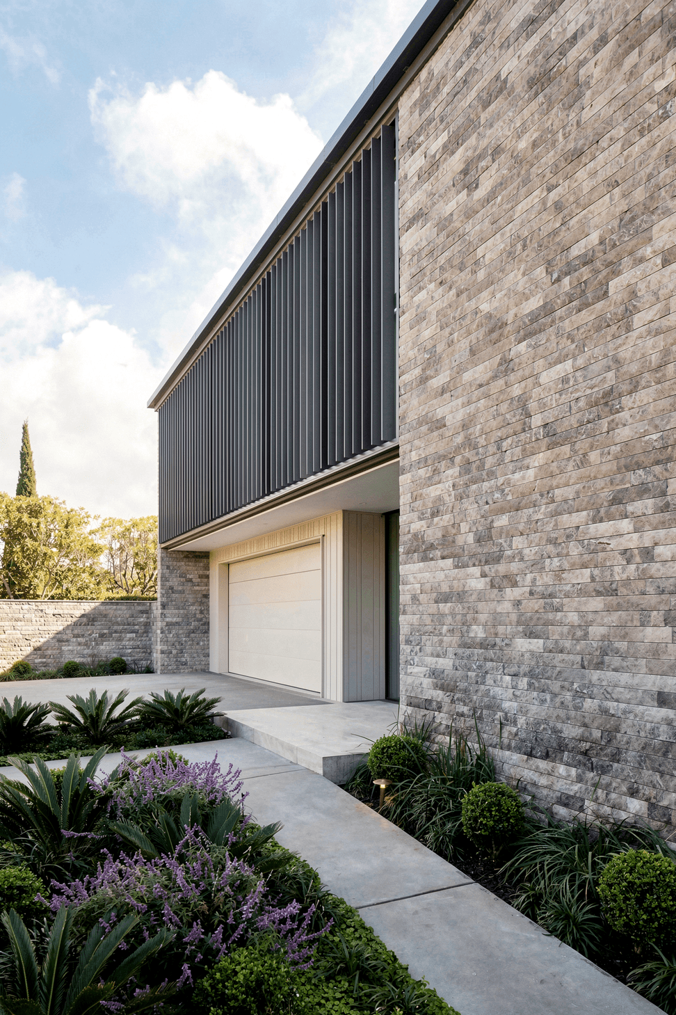 From the front path, the stone-clad corner of the house is softened by lush landscaping. Dark metal louvres introduce texture and privacy, creating a contemporary façade that feels grounded within its garden setting.
