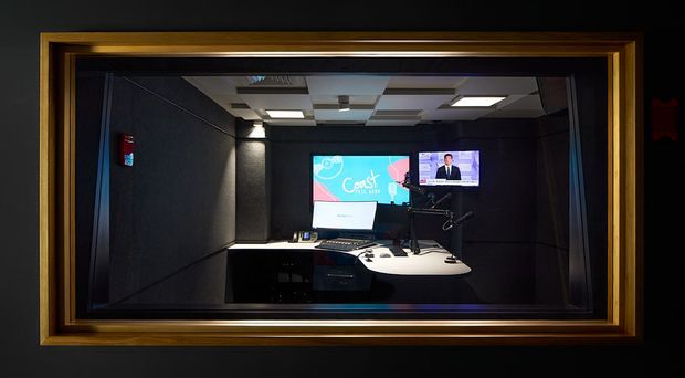 A dark, acoustically treated broadcast or recording studio viewed through a large framed observation window. The room contains a curved control desk with microphones and equipment, multiple screens, and a ceiling with recessed acoustic panels and lighting. The gold-toned timber window frame contrasts with the black wall finishes and subdued studio lighting.