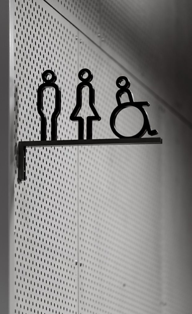 Close-up monochrome photograph of a minimalist accessibility-themed shelf sign, showing male, female, and wheelchair symbols in black metal mounted on a narrow wall shelf. The symbols are set against a perforated light-coloured panel, with strong directional lighting and graphic shadows.
