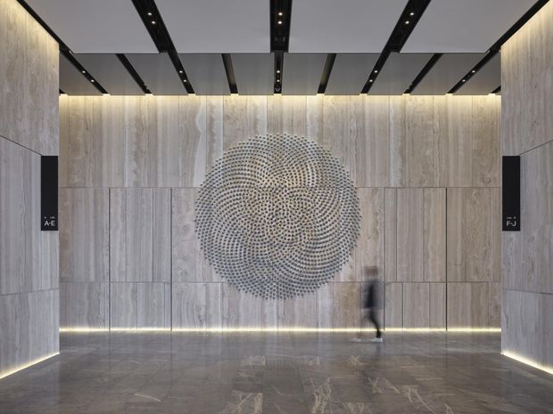 A refined contemporary lobby or entry space with full-height pale stone wall panels, a matching polished stone floor, and concealed linear lighting at the wall base and ceiling edges. A large circular textured metal artwork forms the central feature, while black recessed ceiling beams with integrated spotlights create a strong geometric rhythm. Minimal black wall-mounted signage is visible on both sides, and a blurred person provides scale.
