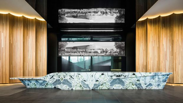 A dramatic contemporary reception lobby featuring a large sculptural reception desk clad in expressive blue, grey and white marbled stone. Warm vertical timber slat walls flank the desk beneath curved scalloped ceiling details, while dark ceilings, reflective glazing and illuminated text panels create a refined, immersive commercial interior.