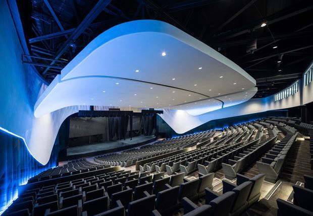 A large contemporary auditorium or lecture theatre with many rows of upholstered seats arranged toward a stage. A dramatic sculptural white suspended ceiling canopy curves above the seating and stage, illuminated by recessed lights and blue accent lighting. The exposed dark structural ceiling and technical stage curtains are visible, creating a high-tech performance or presentation venue.