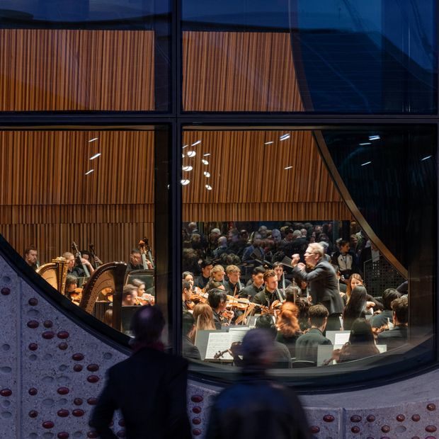 View through large dark-framed glazing into a concert hall, showing a seated orchestra performing beneath warm vertical timber acoustic wall panels. The foreground includes a curved sculptural facade or balustrade with circular red and purple insets, while blurred visitors stand outside the glazing.