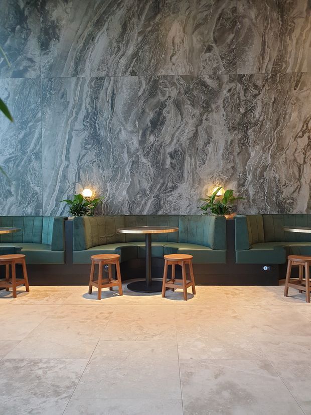 A refined commercial lounge or hospitality seating area with built-in teal and olive upholstered banquettes, small round timber stools, and dark pedestal tables. The backdrop is a dramatic full-height grey veined stone feature wall, complemented by integrated planter niches and warm globe lighting. Large pale stone-look floor tiles create a bright, polished base, while the overall palette is cool, earthy, and sophisticated.