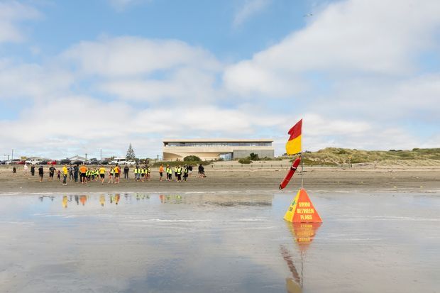 Wide coastal beach scene viewed across wet reflective sand toward a low modern surf lifesaving building and a group of people near the shoreline. A red-and-yellow surf safety flag and warning sign are prominent in the foreground, with dunes and a partly cloudy sky behind.