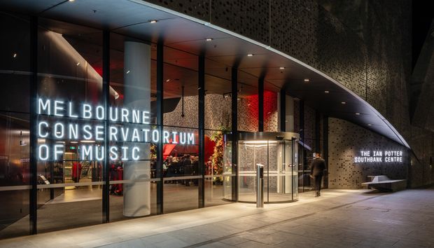 Night-time exterior view of the Melbourne Conservatorium of Music entrance, featuring a curved perforated facade, extensive floor-to-ceiling glazing, illuminated signage, cylindrical revolving doors, and a covered pedestrian forecourt. The interior lobby and reception areas are visible through the glass.