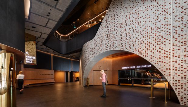 A dramatic contemporary performing arts foyer with a large curved perforated feature wall and arched opening leading to the Kenneth Myer Auditorium. The lobby includes warm timber wall panelling, dark metal finishes, integrated linear lighting, a sculptural black mezzanine or stair enclosure, and polished dark flooring.